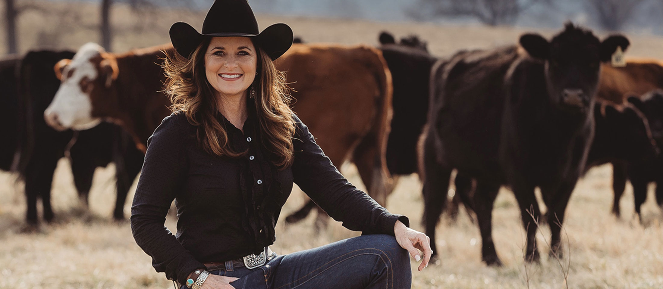 Woman with black hair wearing a black hat and kneeling in a pasture with cattle behind her.